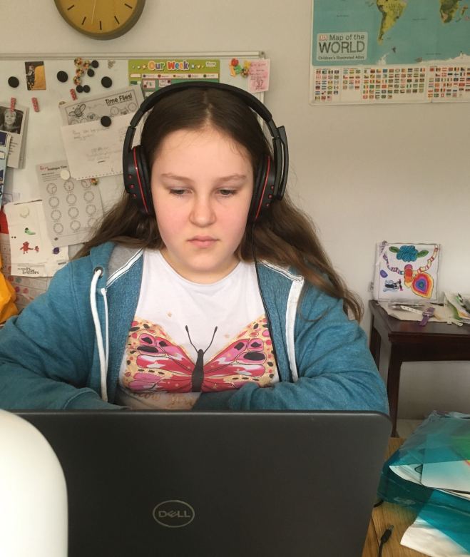 10 year old girl sits facing the camera looking down at an open laptop screen. She is wearing headphones.